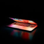 Laptop partially opened, emitting vibrant multicolored lights in dark setting.