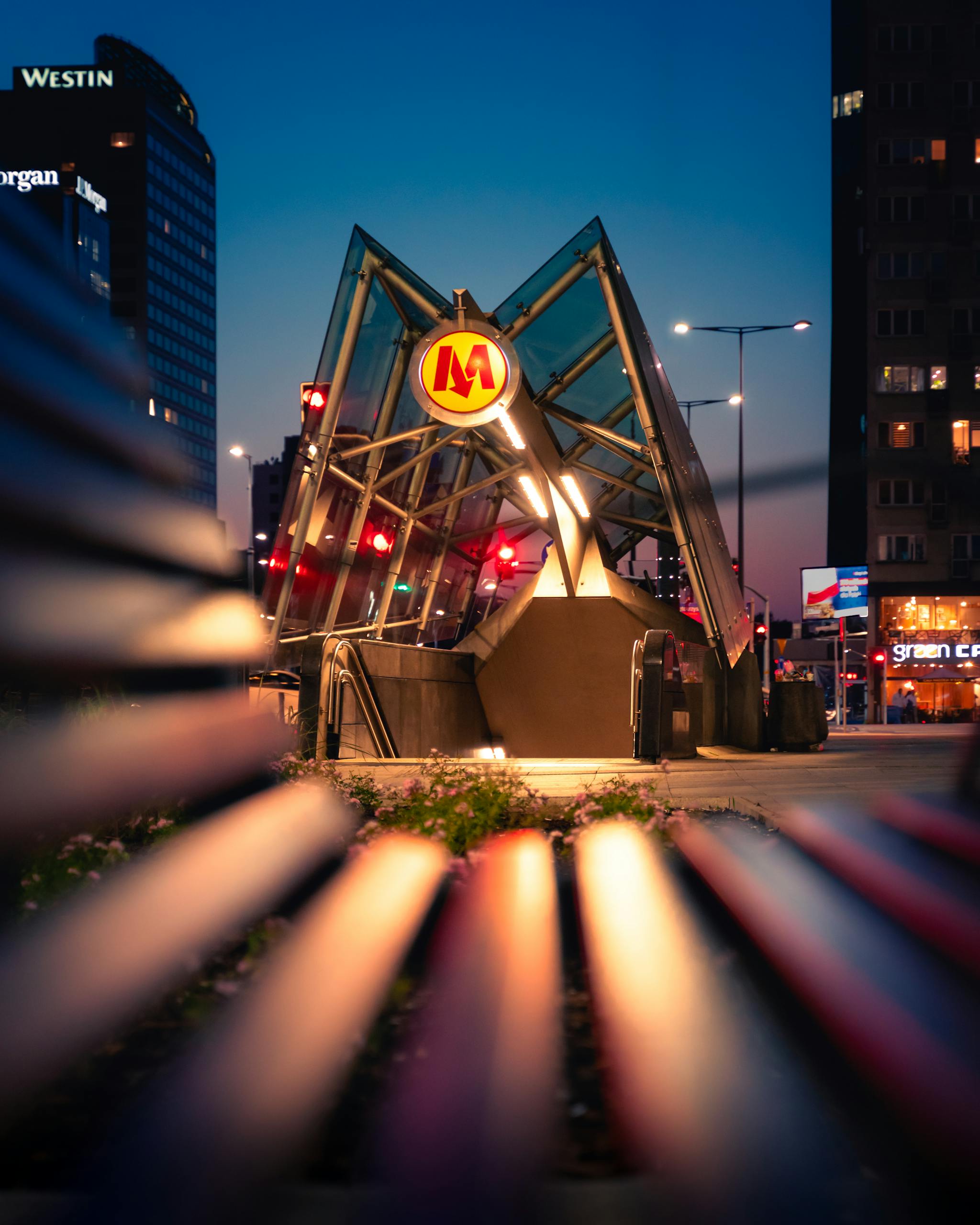 A vibrant view of a Warsaw metro station entrance at dusk, framed by city lights and modern architecture.