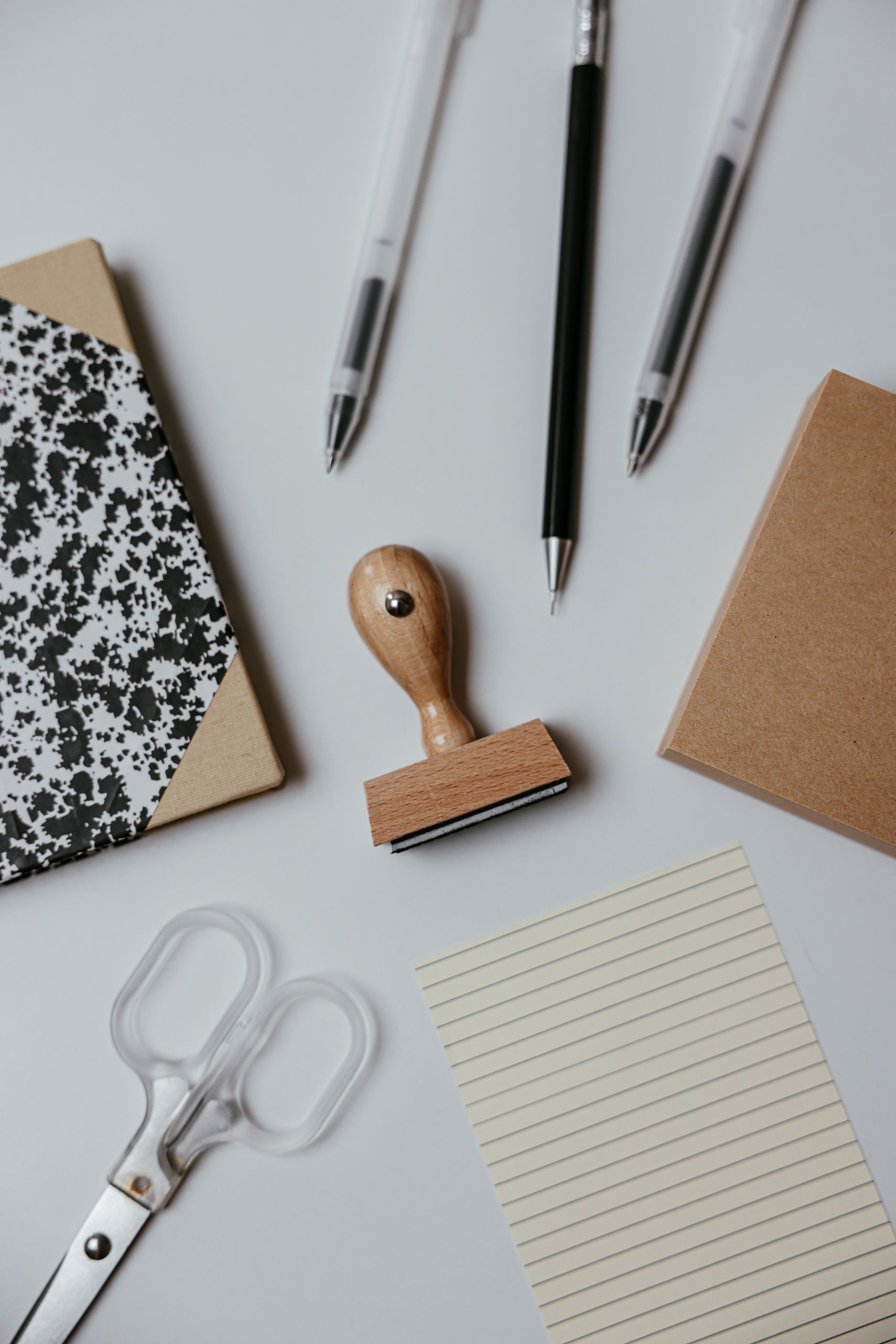 Flat lay of stationery items including notebook, pens, scissors, and stamp on a white surface.
